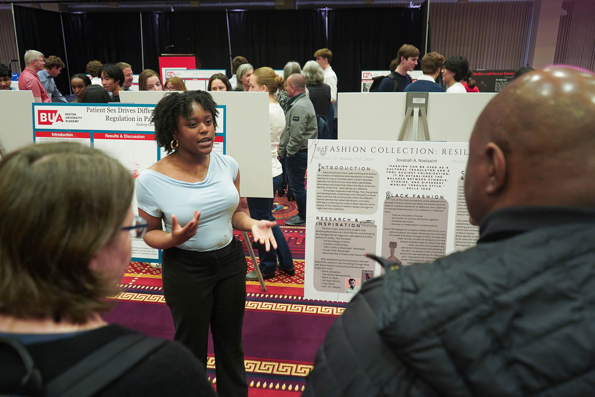Photo: A black woman with short hair in a blue shirt explains a poster board at a recent poster session for thesis students