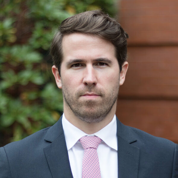Photo: A white man in a suit stands for a formal portrait outside white a stoic expression