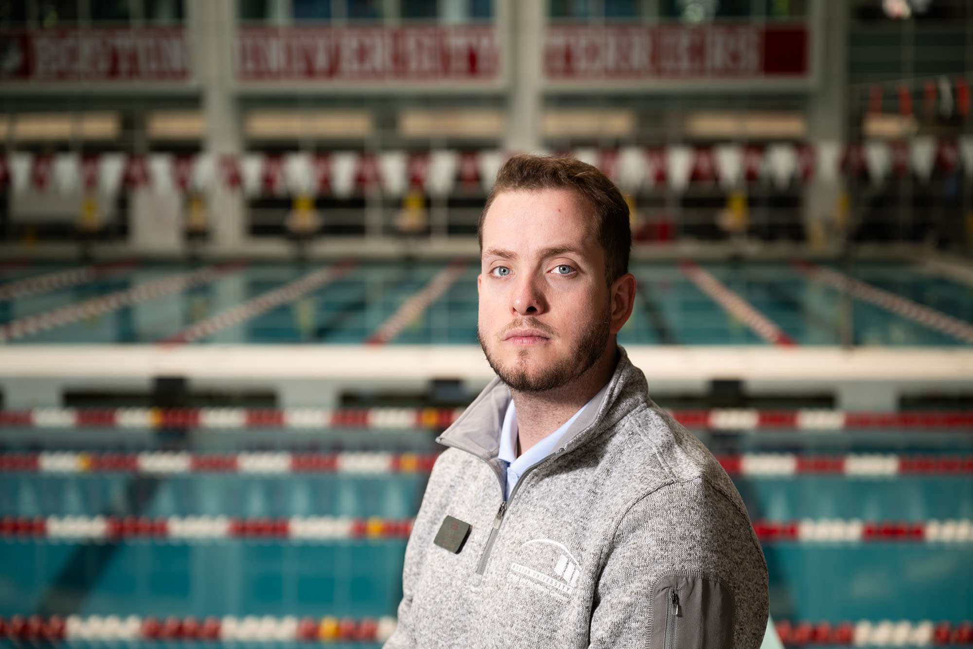 Photo: A picture of a man posing in front of a pool at a university gym. He has short, light brown hair and is wearing a gray zip up