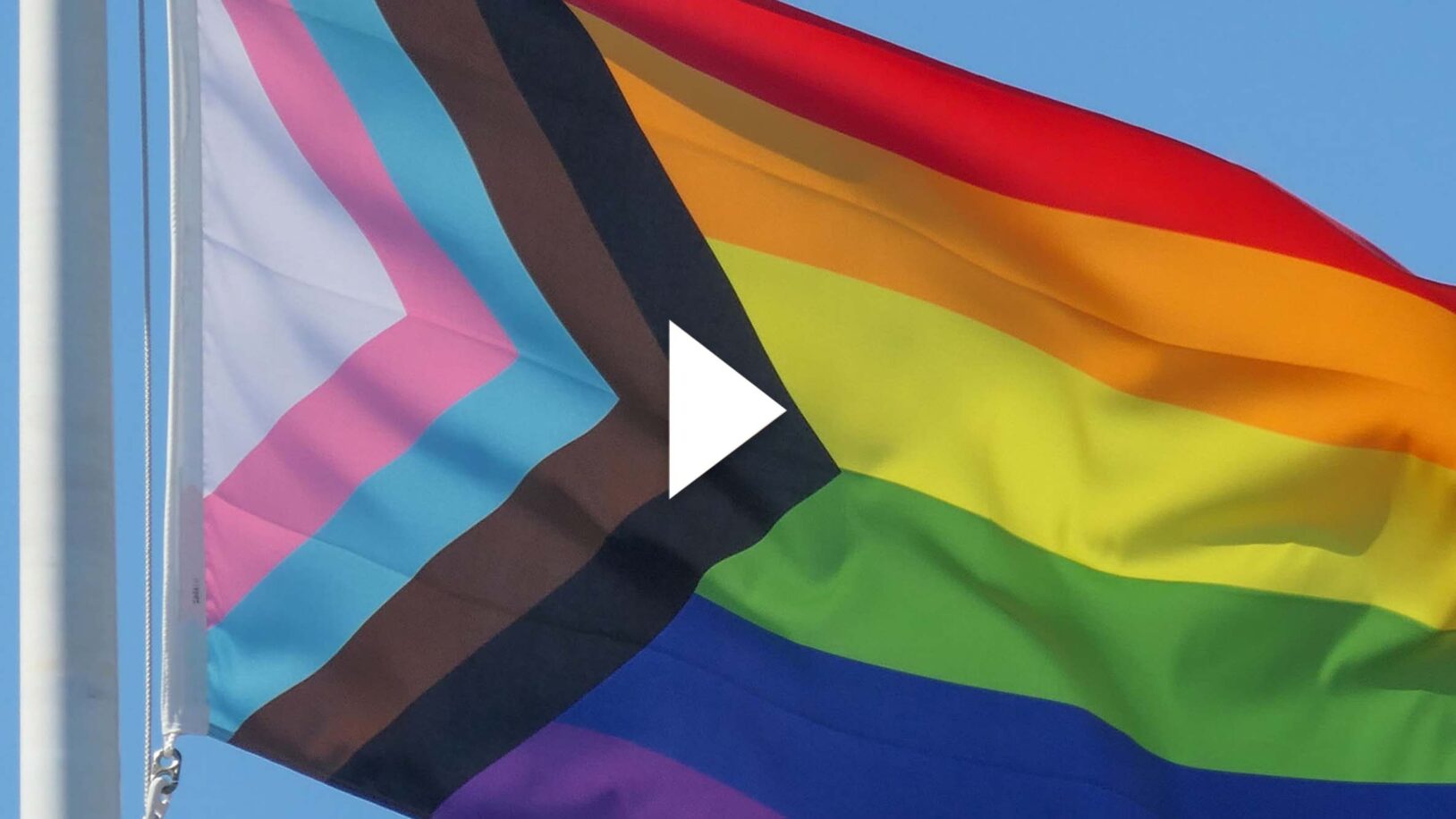 Photo: A picture of a pride flag being raised in front of a blue sky. Play button overlay.