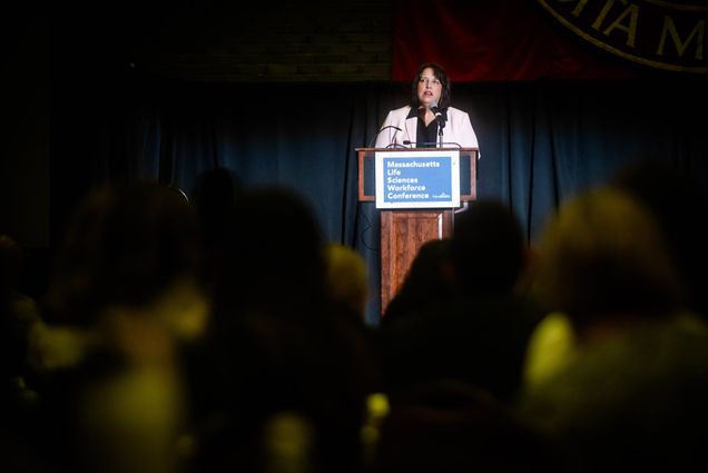 Photo: A white woman, Lt. Gov. Kim Driscoll, stands at a podium addressing a crowd. She wears a white blazer and a black button down.