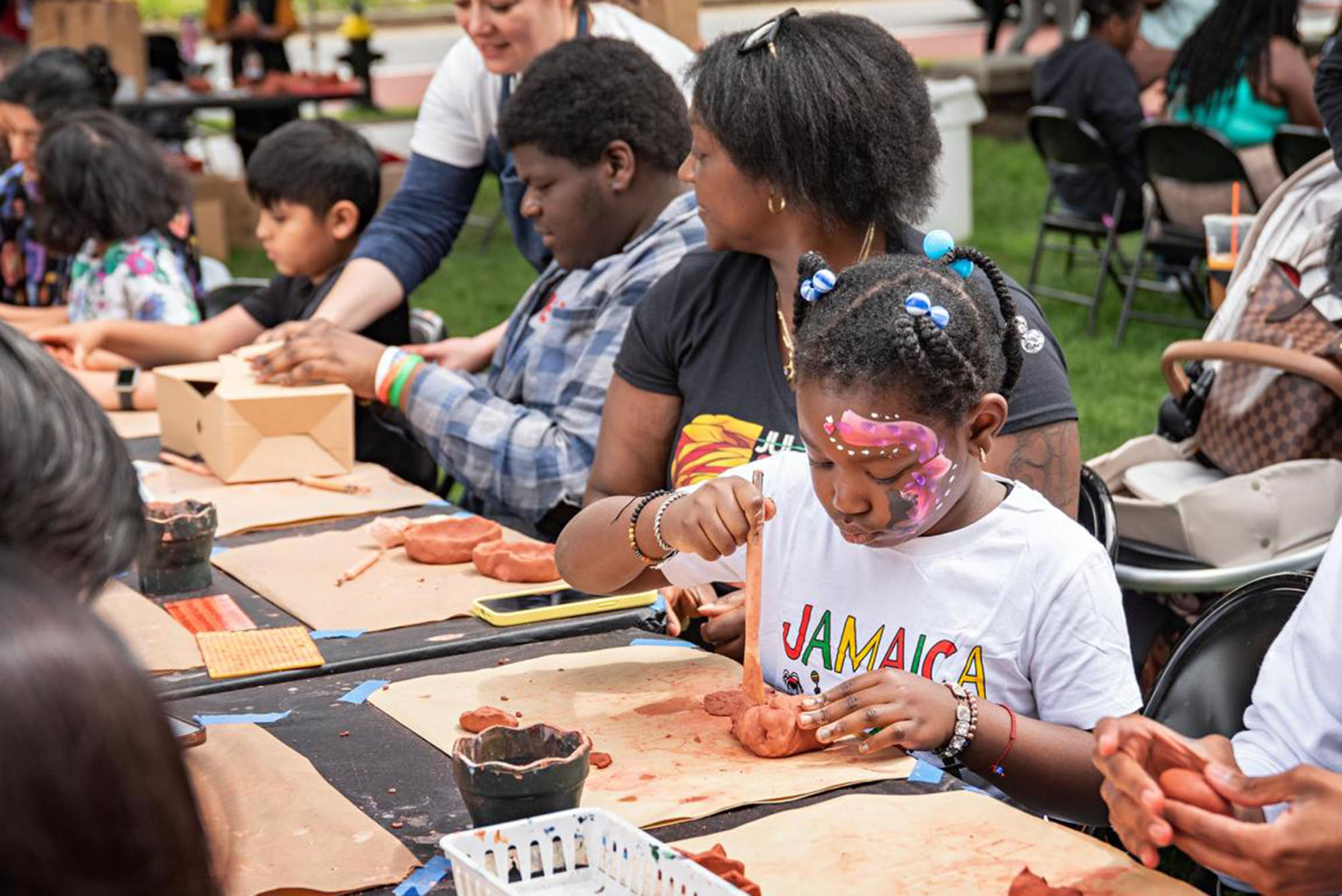 Photo: A picture of an arts and crafts table. At the foreground is a little girl with braids and facepaint painting what appears to be clay 