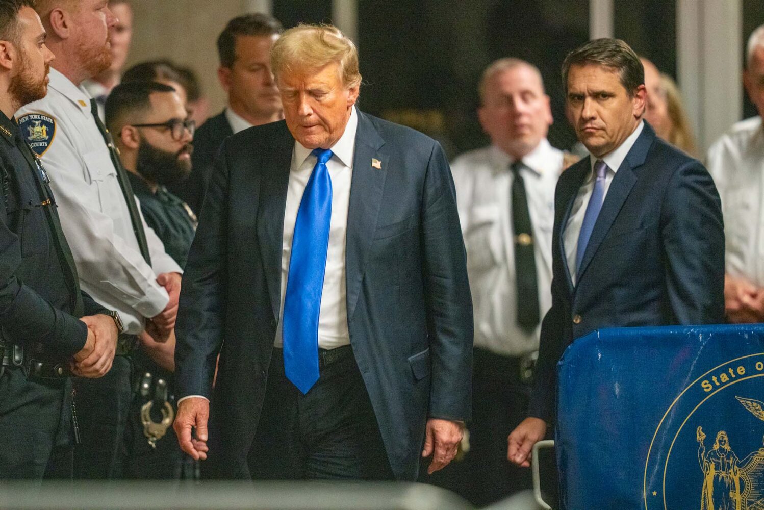 Photo: A picture of former U.S. President Donald Trump walking out of a courthouse. He is wearing a steel blue suit with a bright blue tie and an American flag pin on the jacket