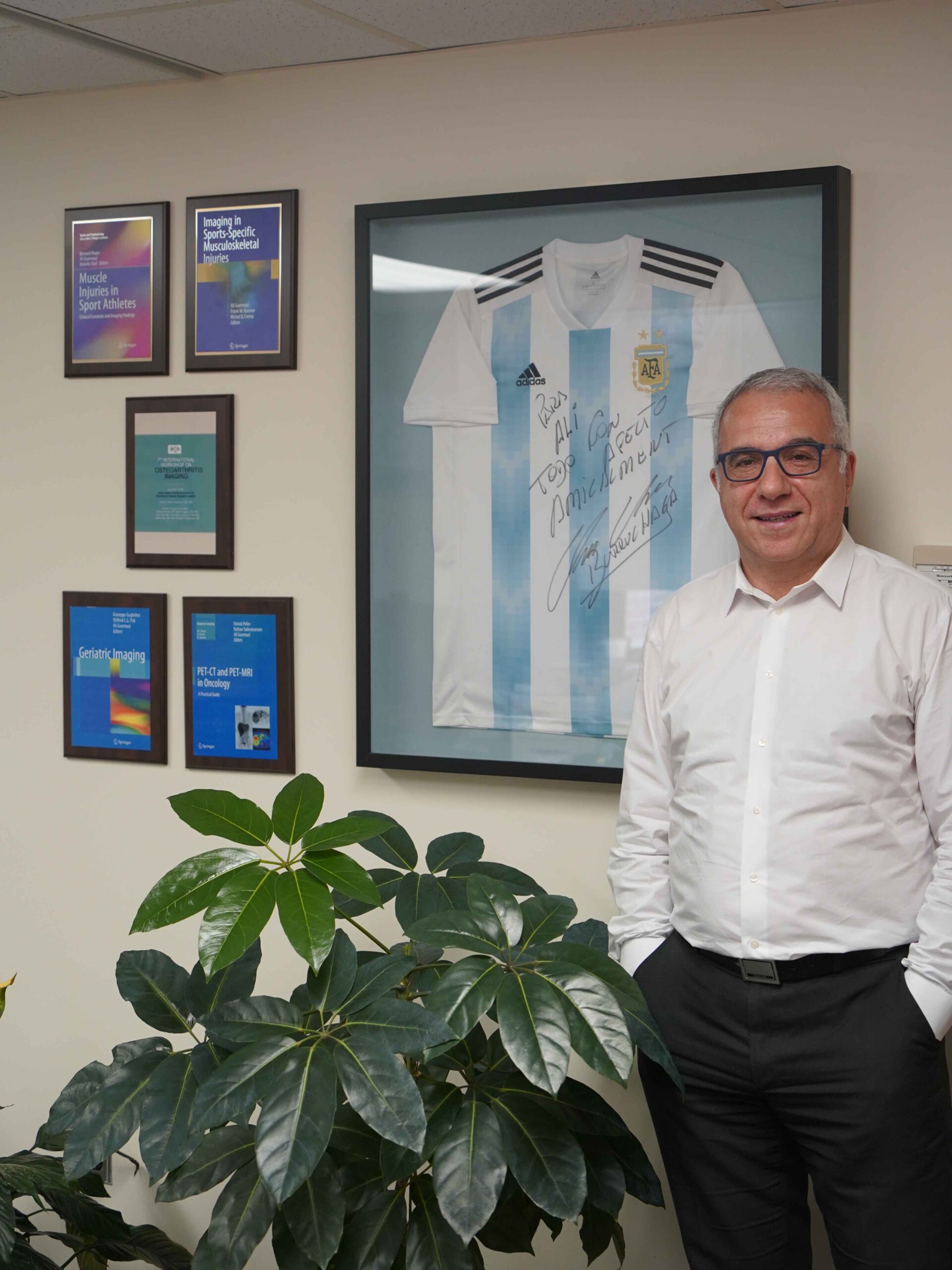 Photo: A picture of a man in a long-sleeve white button down standing in front of a framed, signed soccer jersey