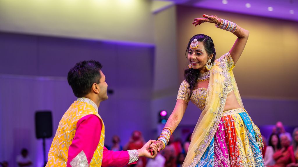 Photo: Two people wearing colorful traditional Indian outfits dancing with each other at an event as onlookers clap