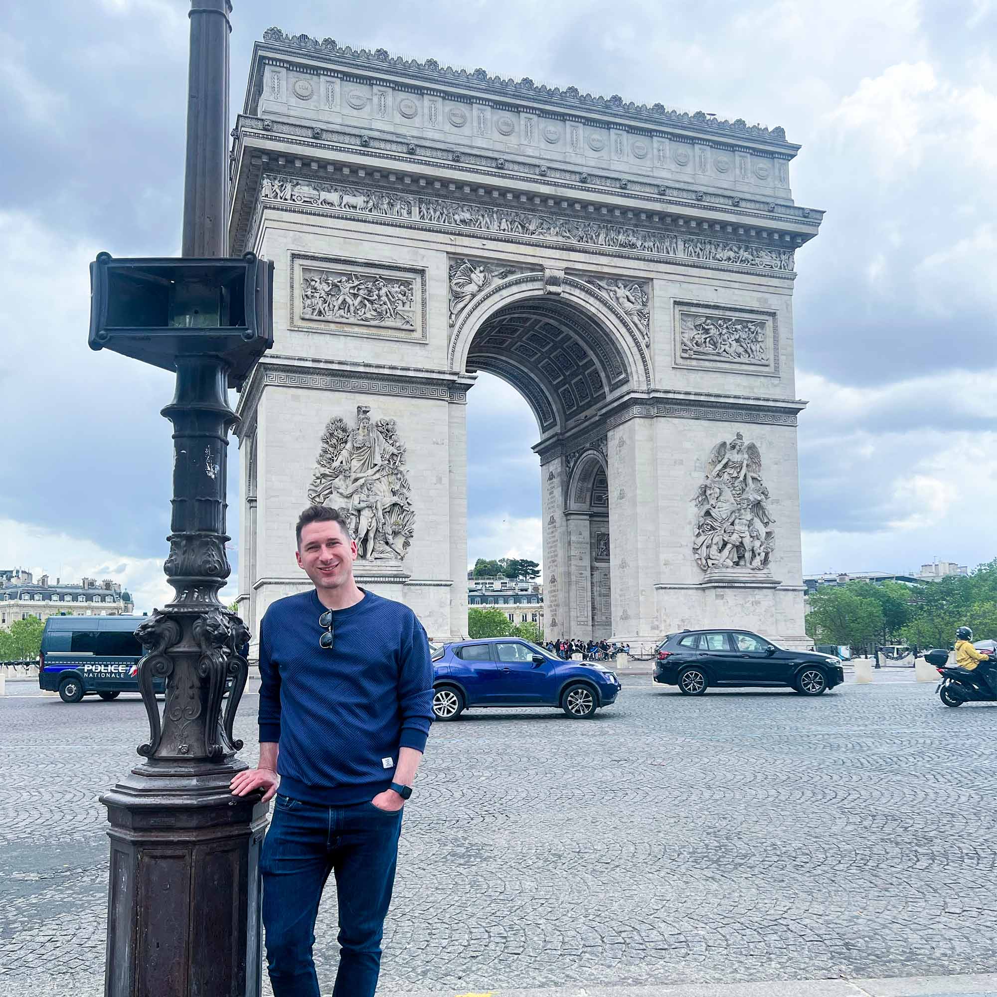 Photo: A young man with dark hair wearing a blue shirt stands in front of the Arc Di Triumph in Paris, France