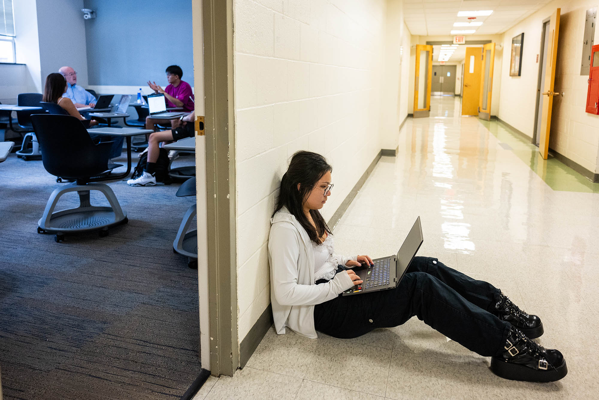 Photo: A young woman sits outside a classroom that is filled with people around a table. She sits on the floor, working on her laptop.