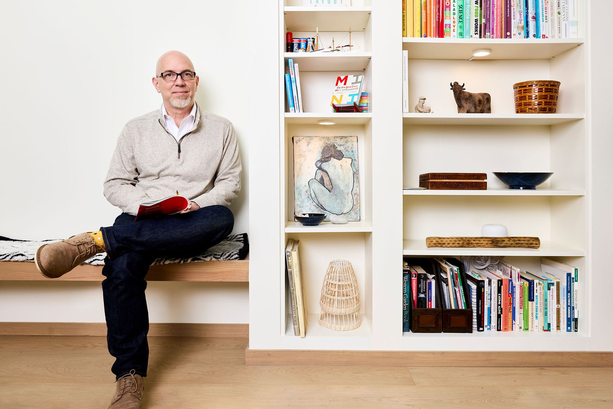 Photo: A bald man with a goatee sits next to a bookcase filled with art and books