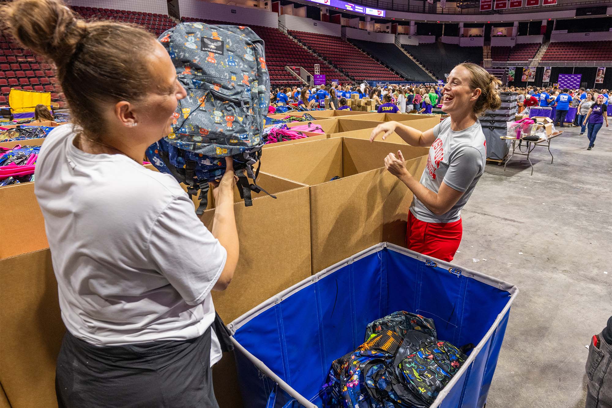 photo: two volunteers standing in a large open room with boxes of backpacks and clothing surrounding them as they sort through