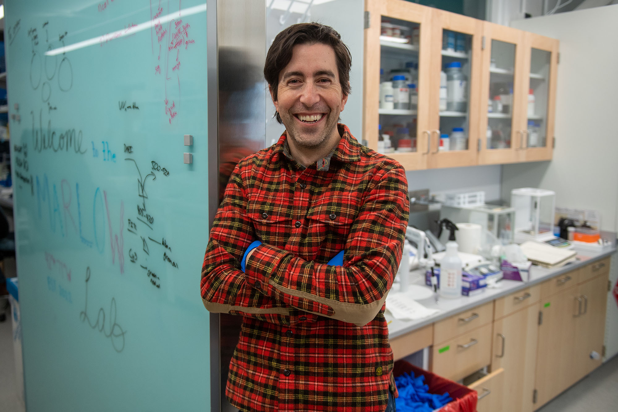 Photo: Jeffrey Marlow, a white man with dark brown hair and wearing a red, black and yellow plaid shirt and blue medical gloves poses in his lab.