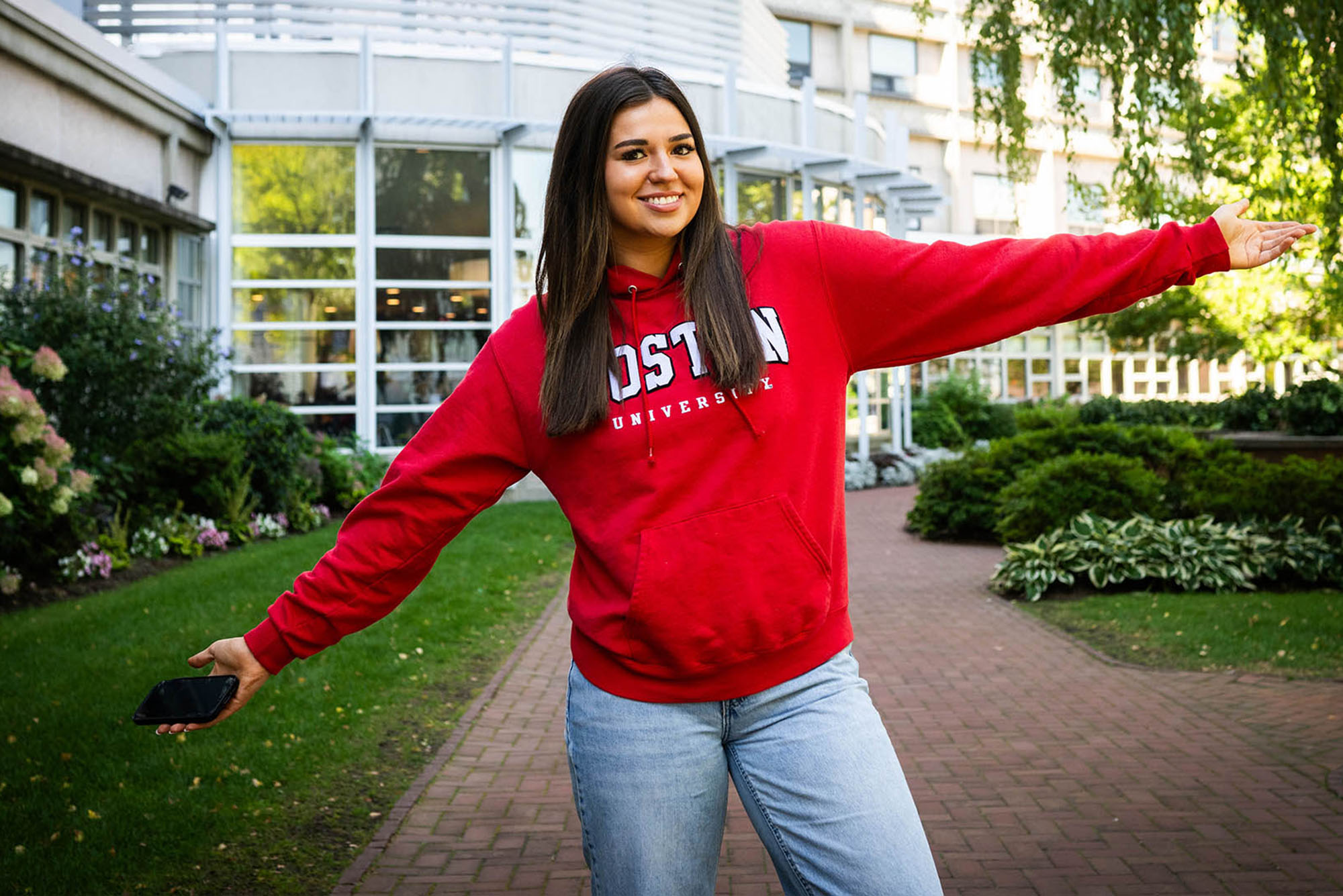 Photo: A woman with long dark hair smiling and posing with her arms extended. She is wearing a bright red sweatshirt that says "Boston University."