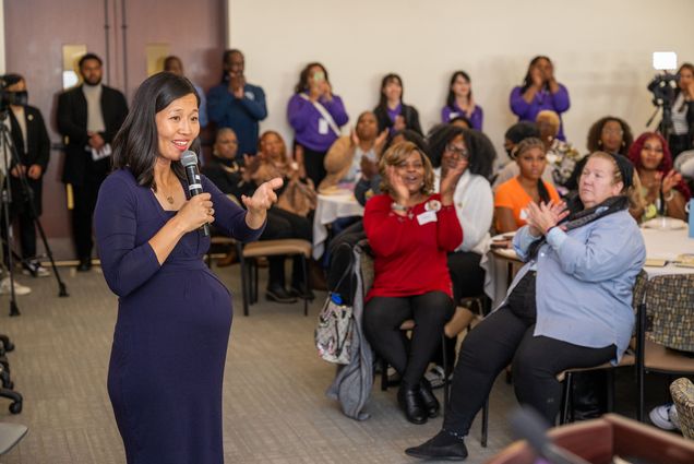 Photo: A picture of Boston Mayor Michelle Wu in a long, indigo dress speaking in front of a group of people. She is standing in front of a seated crowd and holding a microphone