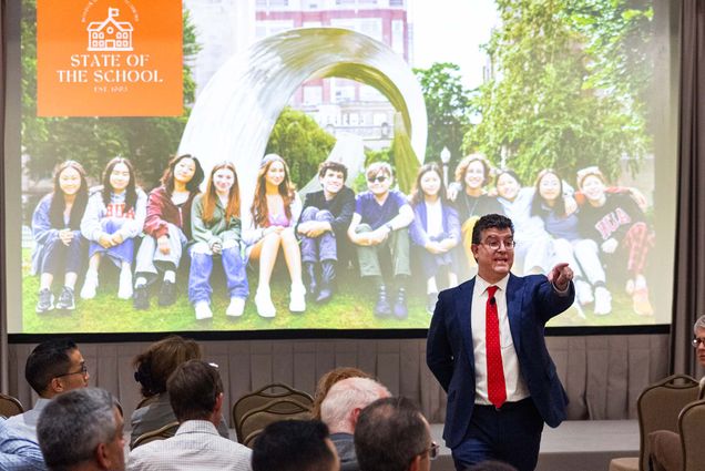 Photo: A man in a suit standing in front of a crowd and pointing towards someone to answer their question. Behind him is a projector screen with a picture of students on it