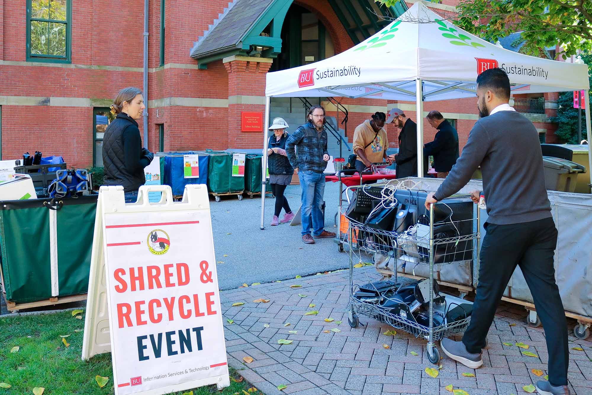 Photo: Tents and bins outside of a red brick building. The tent says "BU Sustainability" and a sign nearby says "Shred & Recycle Event." There are people gathered and a man pushing a cart with electronic equipment towards the tent