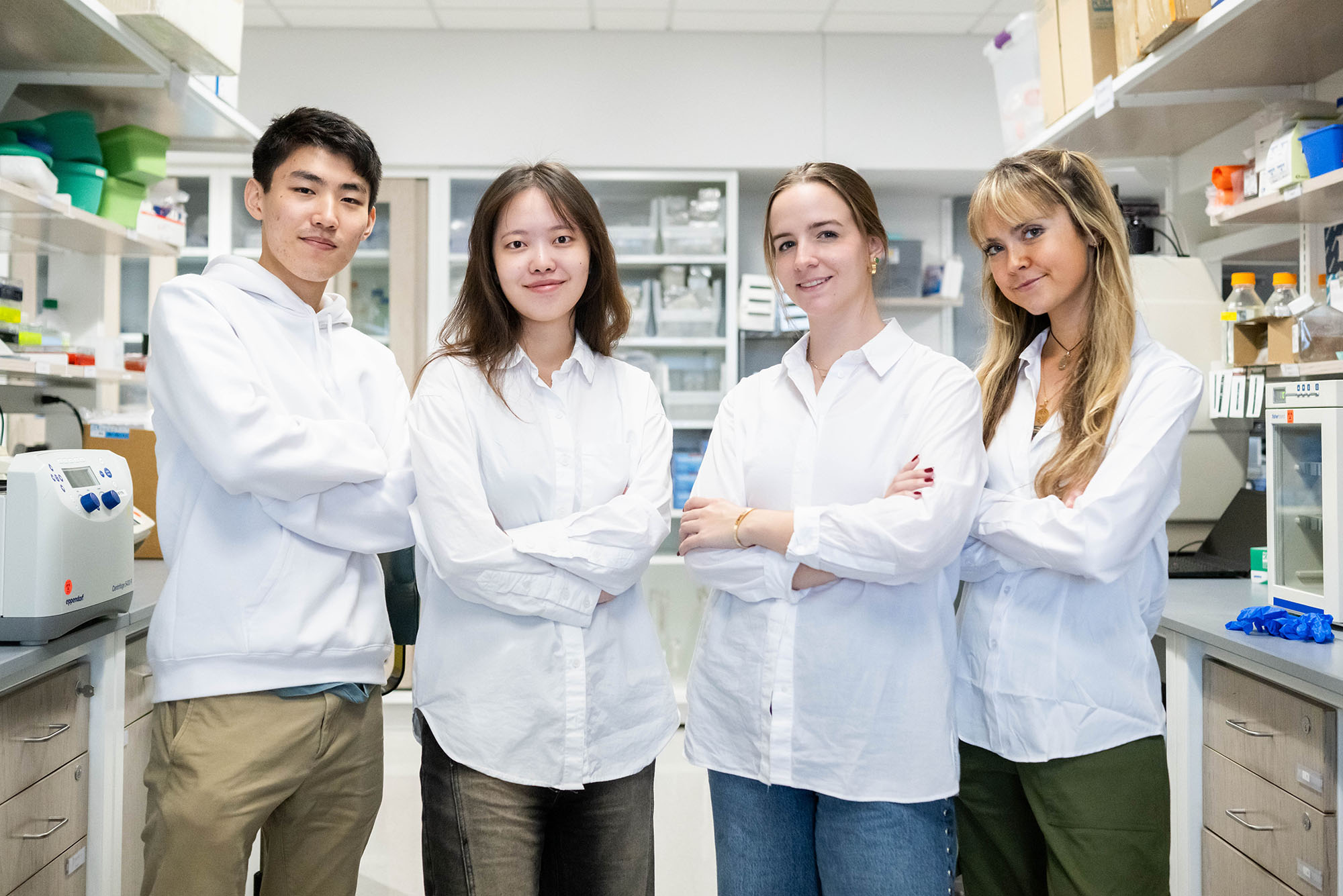 Photo: A picture fo four students in white, long-sleeve shirts with their arms crossed in a biology lab.