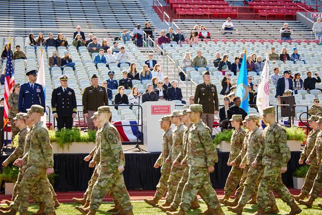 Photo: A picture of people in US Army uniforms marching in front of a stage that has a podium on it with "Boston University" on the front