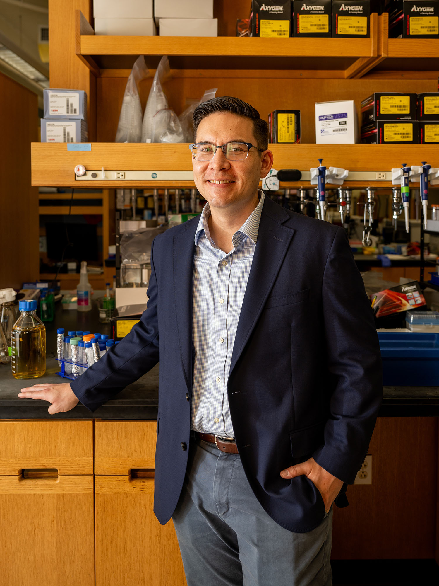 Photo: A picture of a man with short, dark hair and glasses wearing a navy blazer, light blue and white collared button down shirt, a brown belt, and gray pants standing in front of lab equipment.