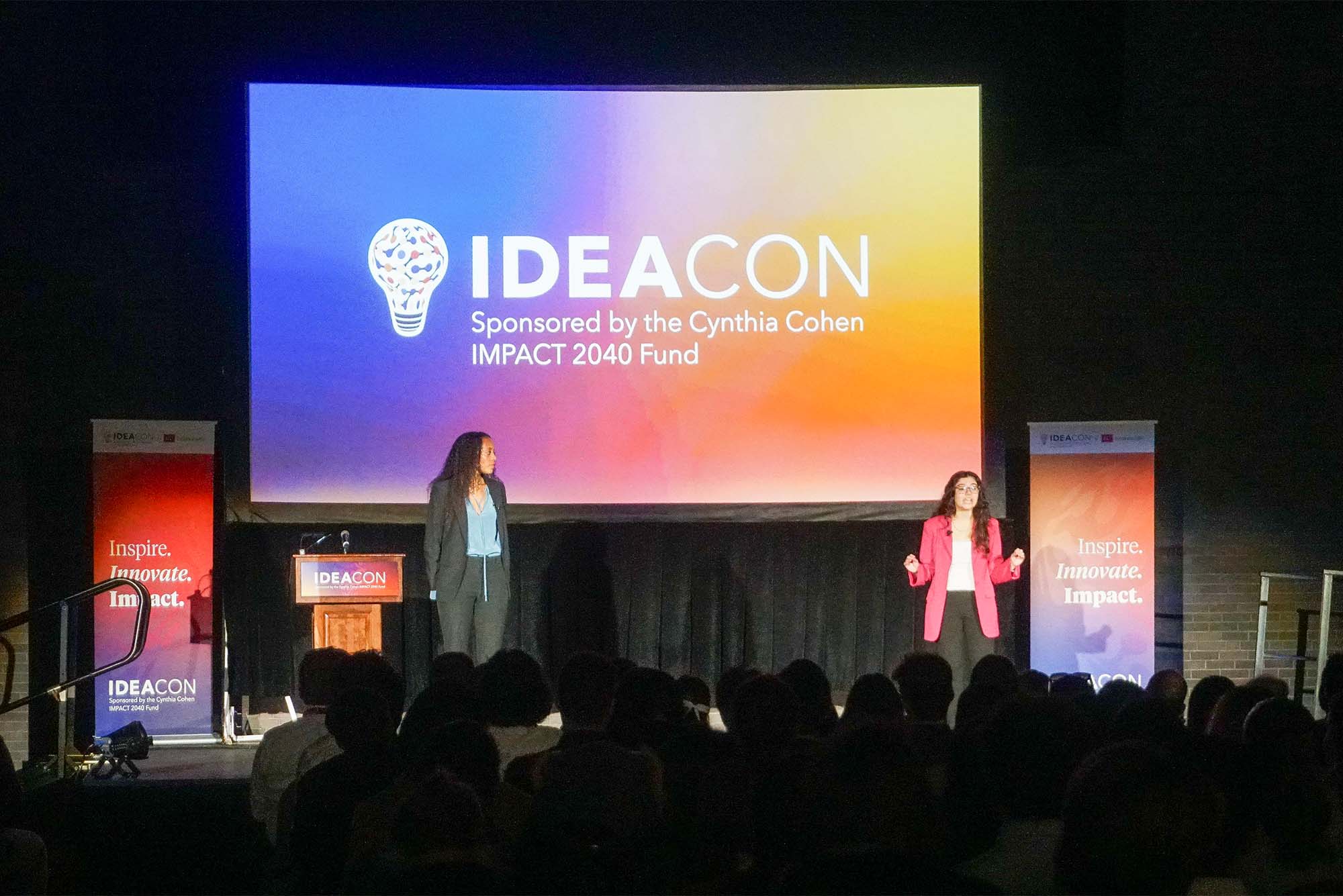 Photo: Two colege student MC's hold microphones under a large blue banner reading "IDEA Con 2024"