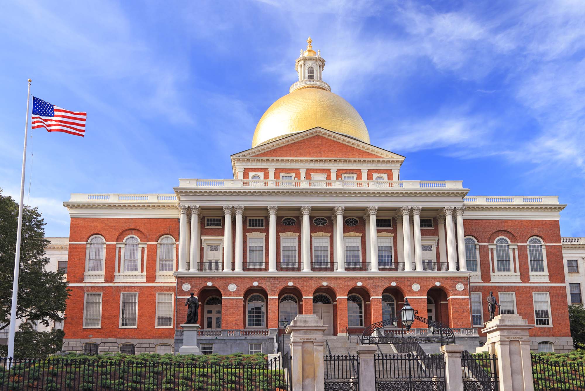 Photo: The Massachusetts State House in Boston, a large red brick building with a golden dome and and an American flag next to it