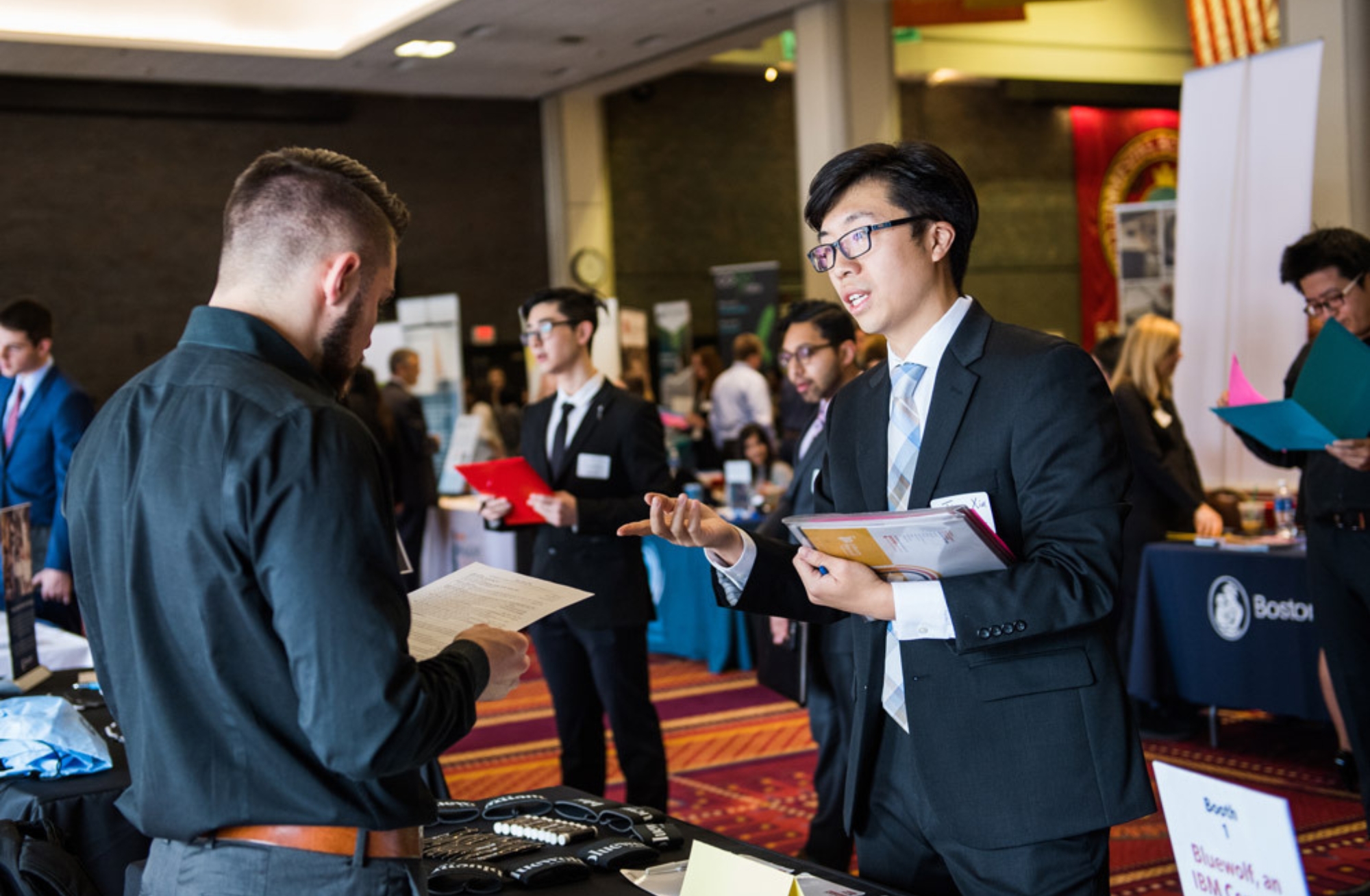 Photo: A busy job fair scene where several people dressed in business attire are conversing. In the foreground, a young man in a suit and tie, holding a folder and documents, is actively speaking with another man wearing a dark shirt who is holding a paper. Other individuals in the background, also in suits, are interacting and exploring the event booths, which have banners and materials set up for recruitment. The event appears to be professional and career-focused, taking place in a large, well-lit room.