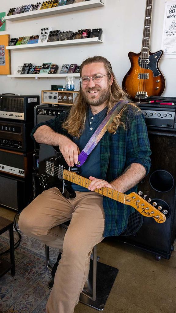 Photo: A picture of a man with long hair wearing a green and blue flannel sittingo n a chair and holding a guitar. He is surrounded by other guitars and guitar pedals