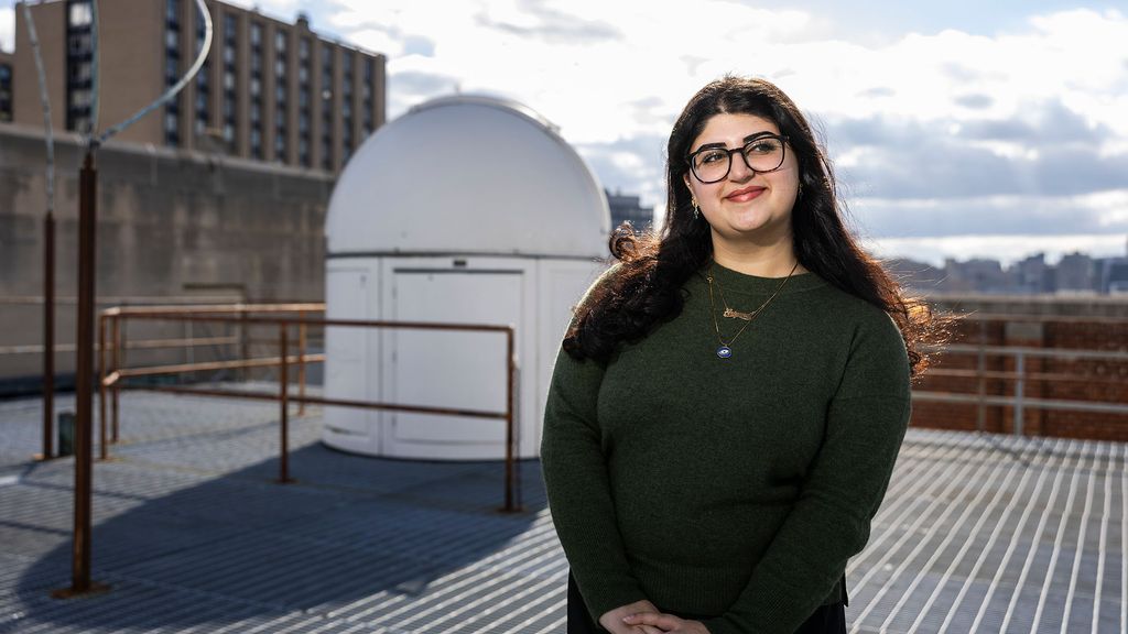 Photo: A female student standing on a rooftop with a closed smile, looking off in the distance
