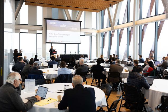 Photo: A large audience sits at round tables to watch a lecturer at a podium give a talk about AI drug discovery