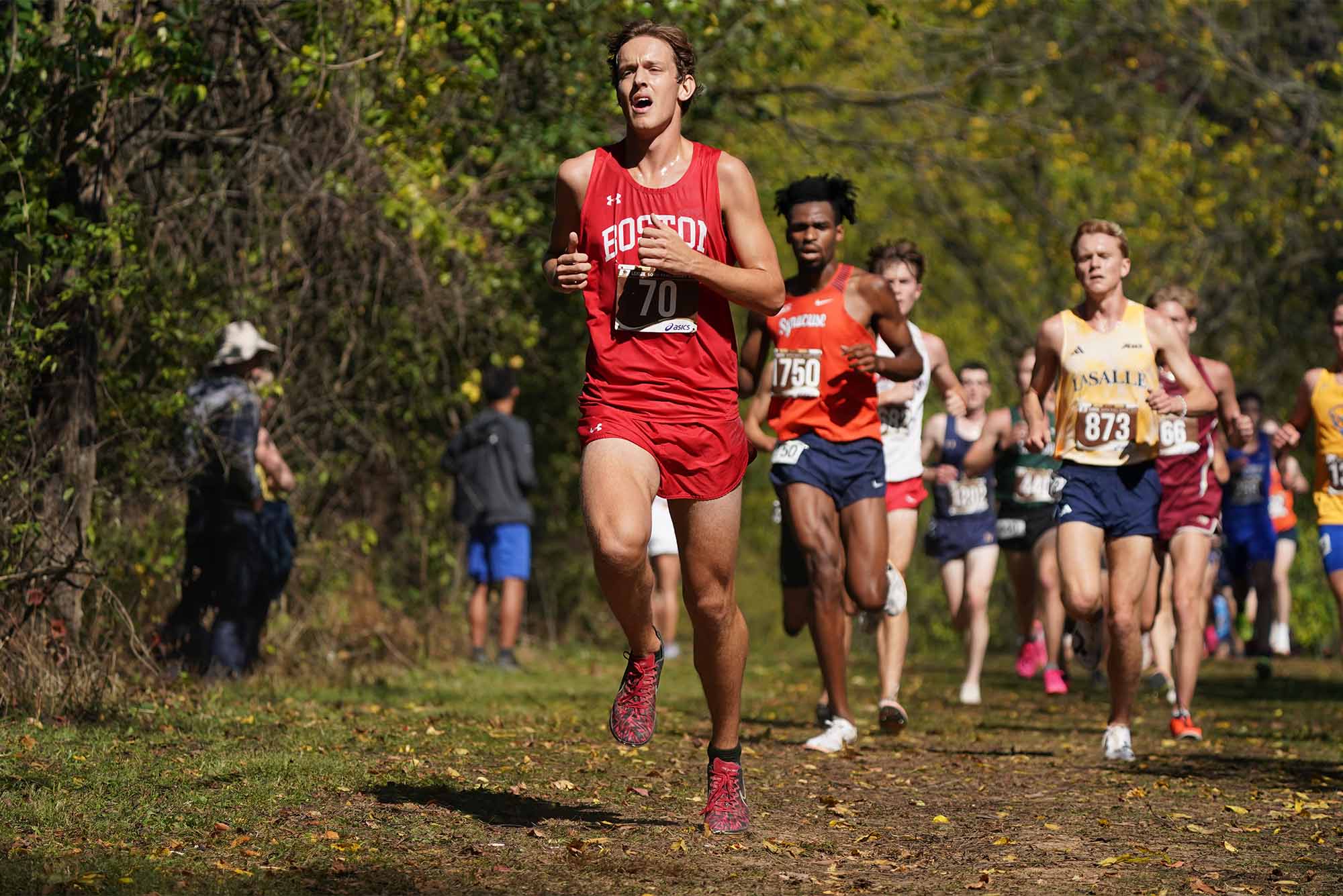 Photo: A men's cross country runner in a red uniform leads a large pack of runners down a trail
