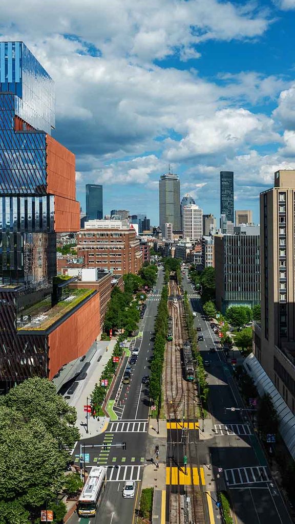 Photo: View of Boston University's urban campus featuring a striking multi-layered glass and metal building, tree-lined streets with tram tracks, and high-rise buildings in the background under a partly cloudy sky.