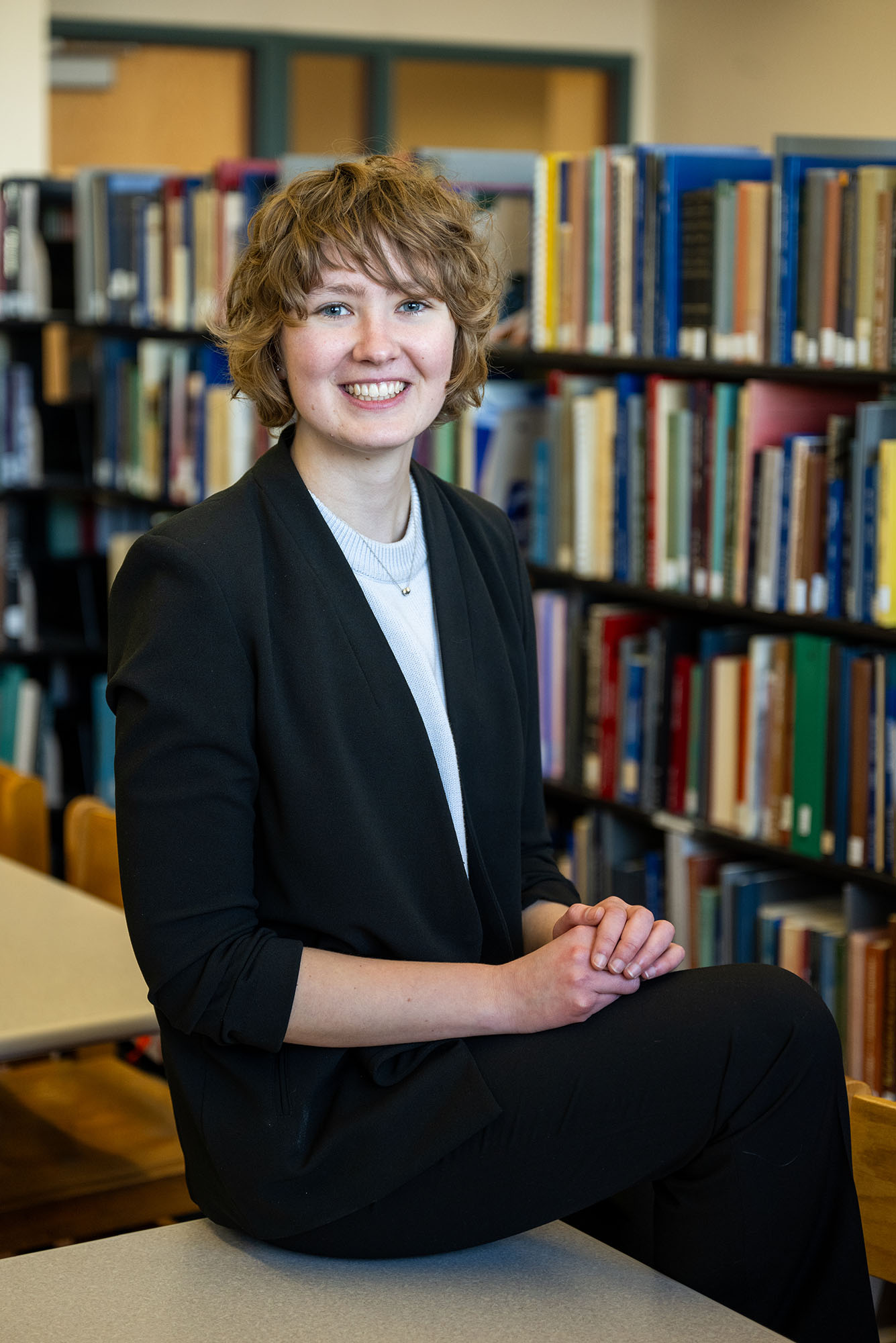Photo: A white woman with short brown hair poses for a portrait. She sits ontop of a table with a bookcase behind her.