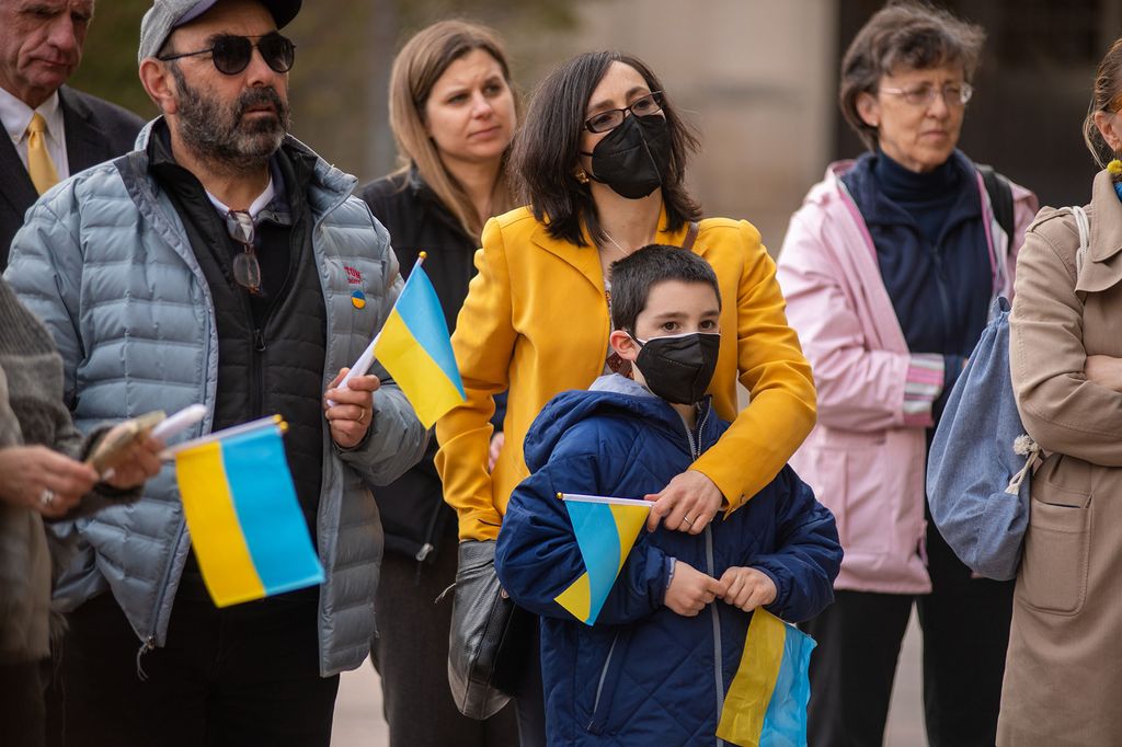 Assistant professor of physics and chemistry, Maria Kamenetska, stands  in the center of the photo wearing a yellow puffer jacket holding her young son, Adam Liston, who is wearing a blue puffer coat. They are both holding a hand-held Ukrainian flag. The flag has a bold, light blue stripe on the top and and a bold, gold-yellow stripe on the bottom. On the left of the pair stands Kamenetska ’s father, Igor Tsinman.  He is wearing a grey baseball cap that matches his grey puffer jacket which is overtop his black zip up. He also has sunglasses on and is carrying the same Ukrainian flag in his left hand. The group stands in front of two women--one in a black coat with light brown hair and the other in a light pink coat with darker and shorter brown hair. They also stand in front of a man in a black suit with a yellow tie, and just behind another woman in a camel coat with a jean draw-string bag. The people around the group are all holding the hand-held Ukrainian flag in their hands.