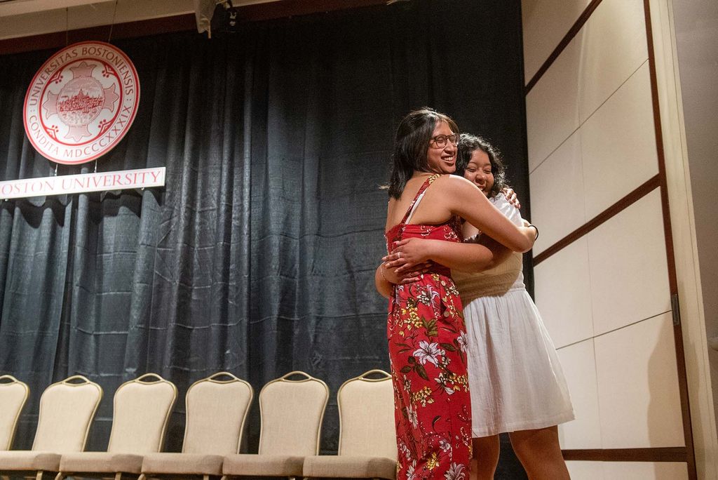 Cecilia Jarquin (SAR’22,SPH’24), in white and on the right, hugging friend, in a red floral jumpsuit on the left. They are at the Newbury Center’s First Gen Pinning Ceremony at Questrom, standing on top of the stage. Behind them are empty beige chairs, a large BU crest, and a grey-ish blue curtain. The two are smiling. 