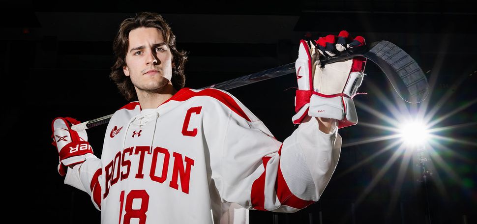 Photo: A hockey player posing in his Boston University uniform with a hockey stick in front of a black background
