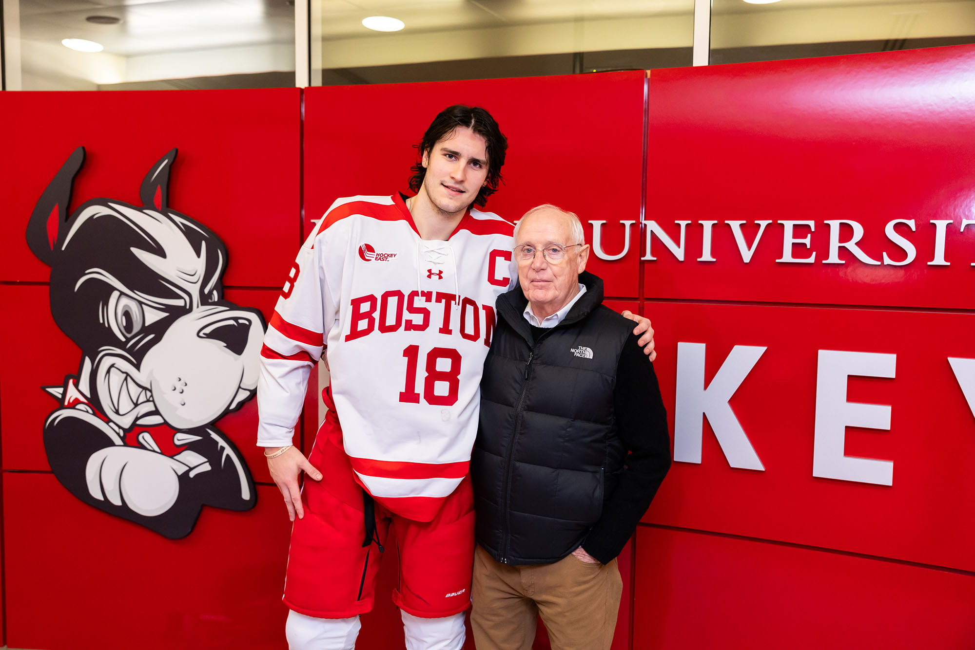 Photo: A hockey player in Boston University uniform on the left with his arm around a man with white hair wearing a North Face vest on the right