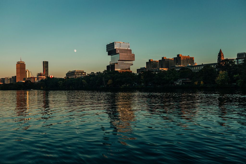 Photo: A wide shot of a river and and a skyline. It is dusk out, with the moon on the left-hand side, full. In the center of the skyline stands a building, rectangular save for the top, which juts out to the right.