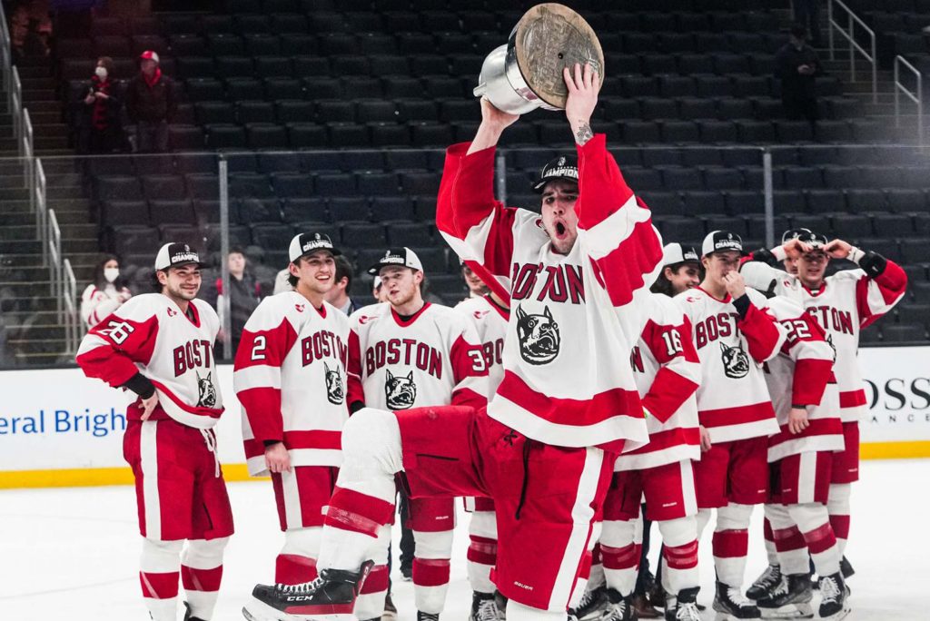 Photo of Dylan Peterson holding the Beanpot trophy after BU beat Northeastern during the 69th Annual Beanpot tournament championship game on February 14 at TD Garden. The male hockey player is caught mid-jump as he hold a silver trophy above his friends. His teammates smile and look on behind him on the hockey rink
