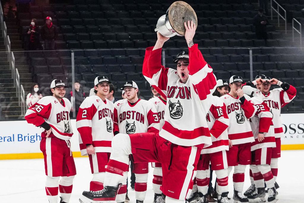 Photo of Dylan Peterson holding the Beanpot trophy after BU beat Northeastern during the 69th Annual Beanpot tournament championship game on February 14 at TD Garden. The male hockey player is caught mid-jump as he hold a silver trophy above his friends. His teammates smile and look on behind him on the hockey rink