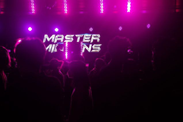 Photo: A dim-lit room, illuminated by fluorescent, pink neon lights. A crowd is at the foreground of the photo while in the background, a DJ stand and DJ set the vibes. The DJ is off-center, toward the left. A huge sign behind the DJ reads "MASTER" and unreadable second word.