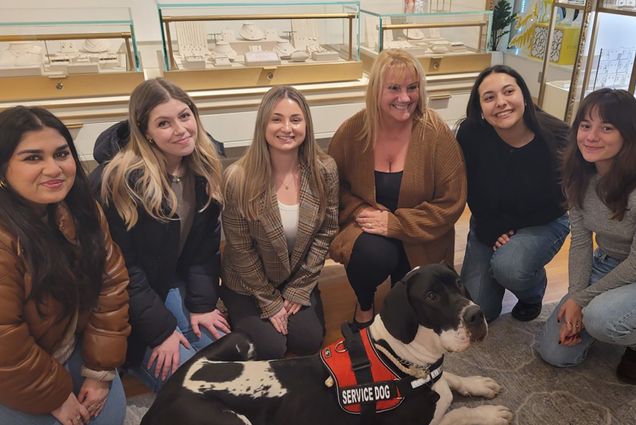 Photo: A group of woman sit in the center of the frame, surrounding a black and white dog. From the left, the woman has dark hair and a brown puffer coat, the next a black puffer coat and dirty blonde hair, the next a beige cardigan and blonde hair, the next a darker brown cardigan with blonde hair, the next a brunette wearing a black long sleeve shirt, and the last woman has on a grey shirt with dark brown hair. The dog has on a red vest indicating that the dog is a service dog.