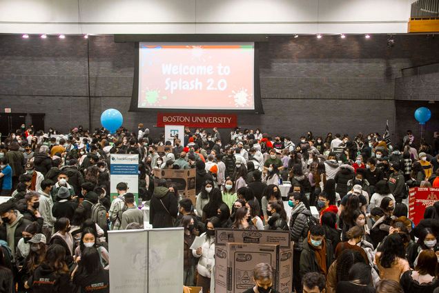 Photo: A large crowd of students in an auditorium -type arena. The area is gray and monotoned. Behind them is a projector screen displaying the words, in white bubbly font, "Welcome to Splash 2.0!". The background is red.