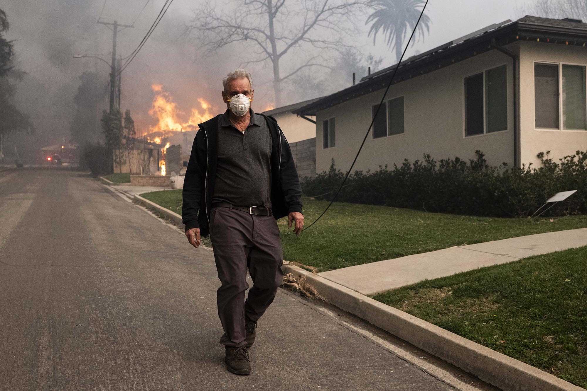 Photo: A man with a mask walks on the road, flames and smoke blazing behind him.