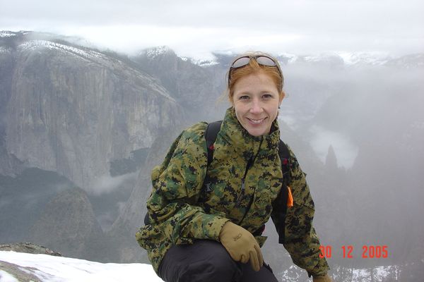 Photo: A woman with red hair smiling and posing in front of a snowy mountain landscape while wearing a camo jacket