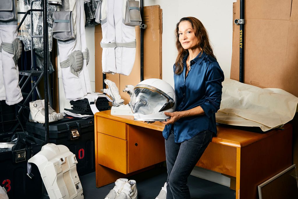 Photo: A woman leaning against a desk as she holds an astronaut helmet. The wall behind her has astronaut suits across it and a poster with an astronaut that says "Apply Now"