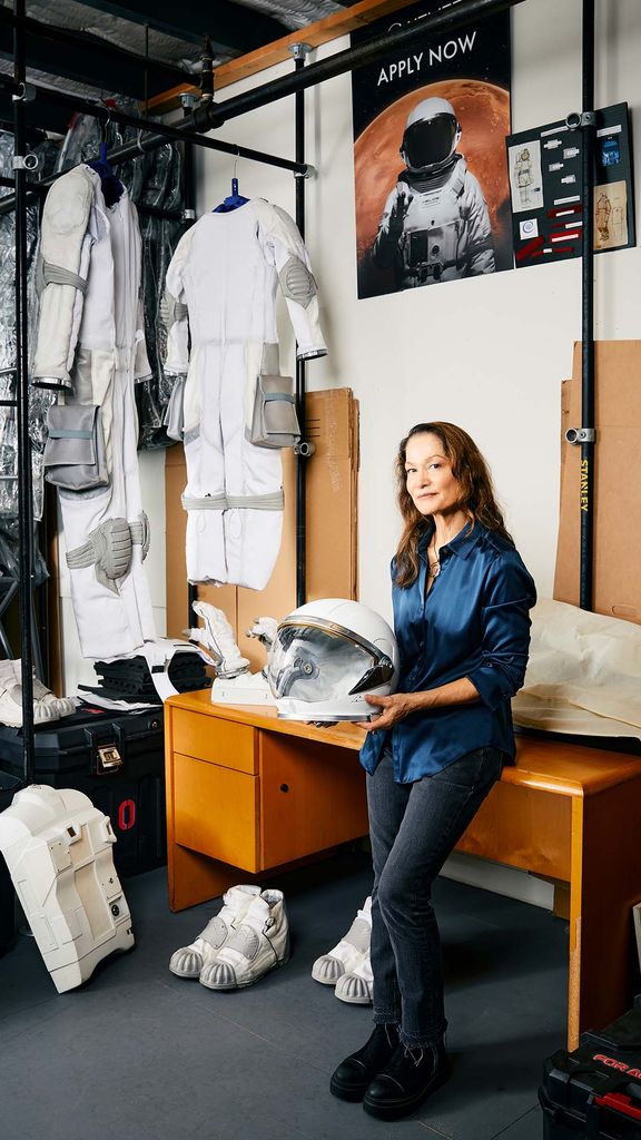 Photo: A woman leaning against a desk as she holds an astronaut helmet. The wall behind her has astronaut suits across it and a poster with an astronaut that says "Apply Now"