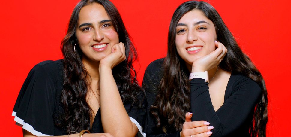 Photo: Two girls smiling and posing with their head resting against their hand as they sit behind a table. There is a red background behind them