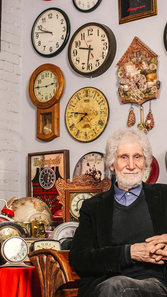 Photo: A distinguished man with white hair sits with hands folded in front of a wall of ornate clocks and other items
