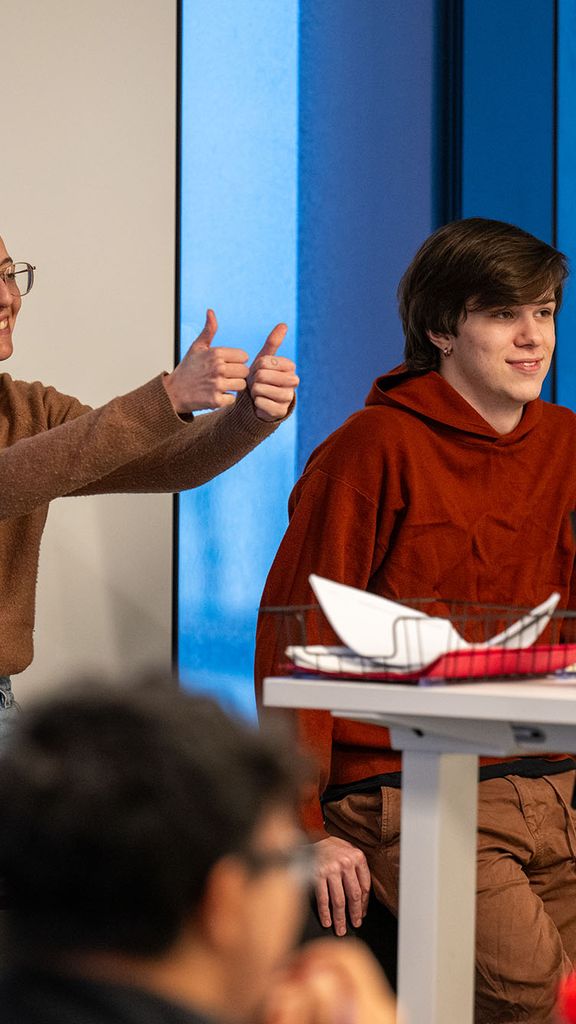 Photo: Two people sitting in front of a larger group, one on the left is giving a thumbs up and the other sits in front of a computer