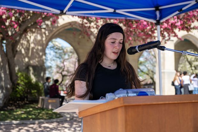 Photo: A girl standing behind a podium and reading outside with crab apple trees in the background
