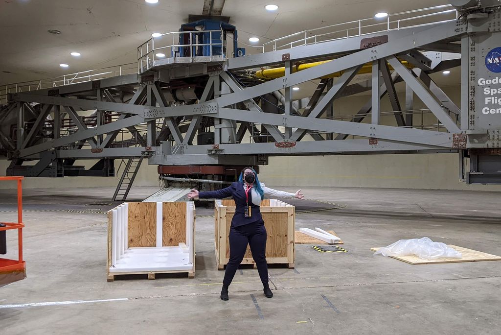 Photo: A person with long, blue hair wearing a mask and spreading outu their arms in a NASA center
