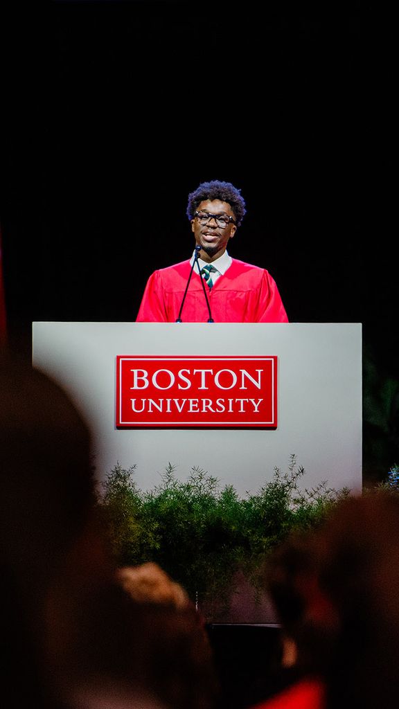 Photo: BU Student Government President Akwasi Antwi talking behind a podium that says "Boston University" while wearing a red academic gown